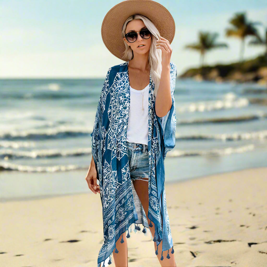Woman wearing a patterned boho kimono-style cover-up in blue, paired with a wide-brimmed hat, white top, and denim shorts. Smaller images feature the same cover-up in black, teal, and rust colors—elegant, flowy, and perfect for layering.