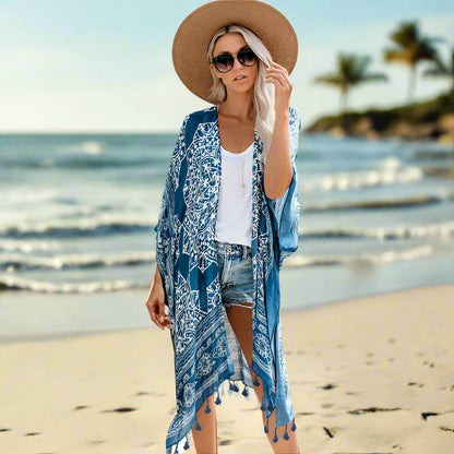 Woman wearing a patterned boho kimono-style cover-up in blue, paired with a wide-brimmed hat, white top, and denim shorts. Smaller images feature the same cover-up in black, teal, and rust colors—elegant, flowy, and perfect for layering.