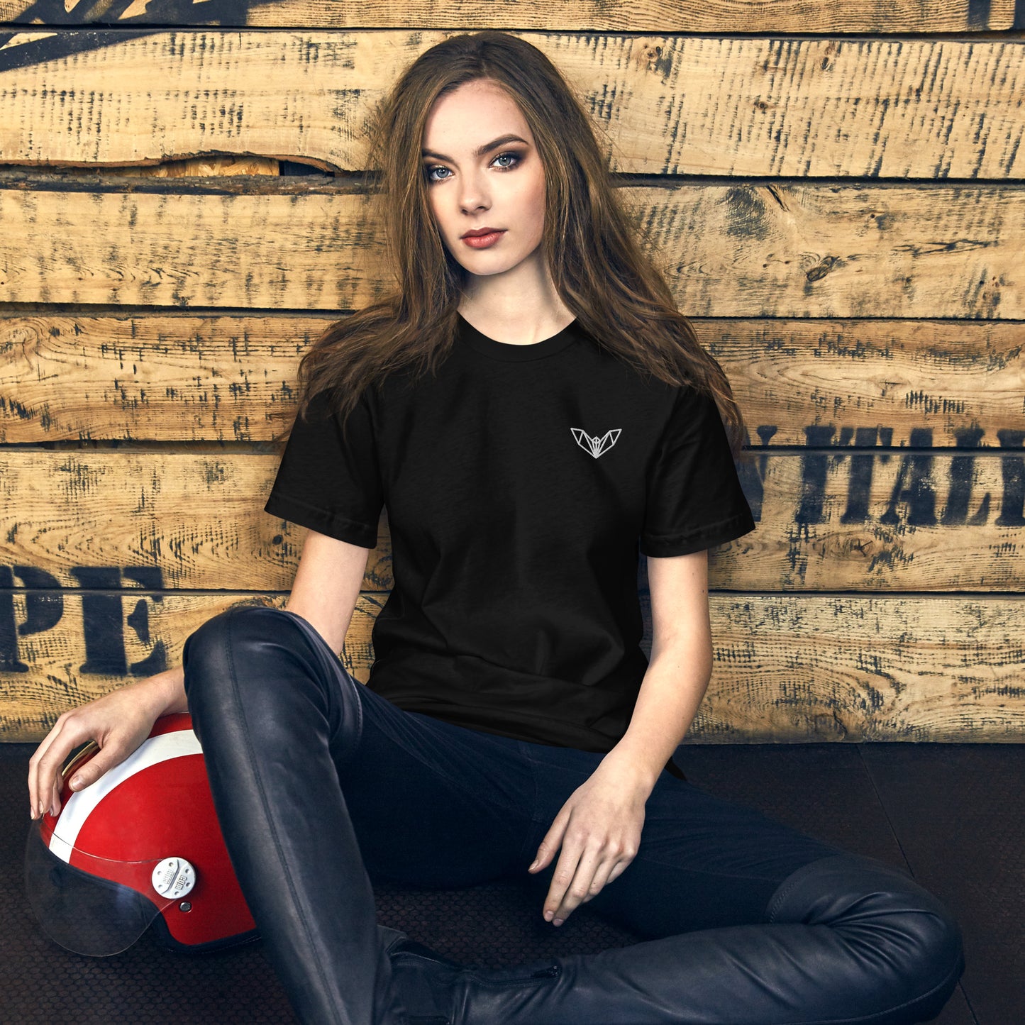 Black women’s t-shirt featuring a small white geometric bat logo on the left side of the chest, worn while seated on a wooden floor with a red and white helmet nearby—sleek, minimalist, and perfect for casual and athletic wear.