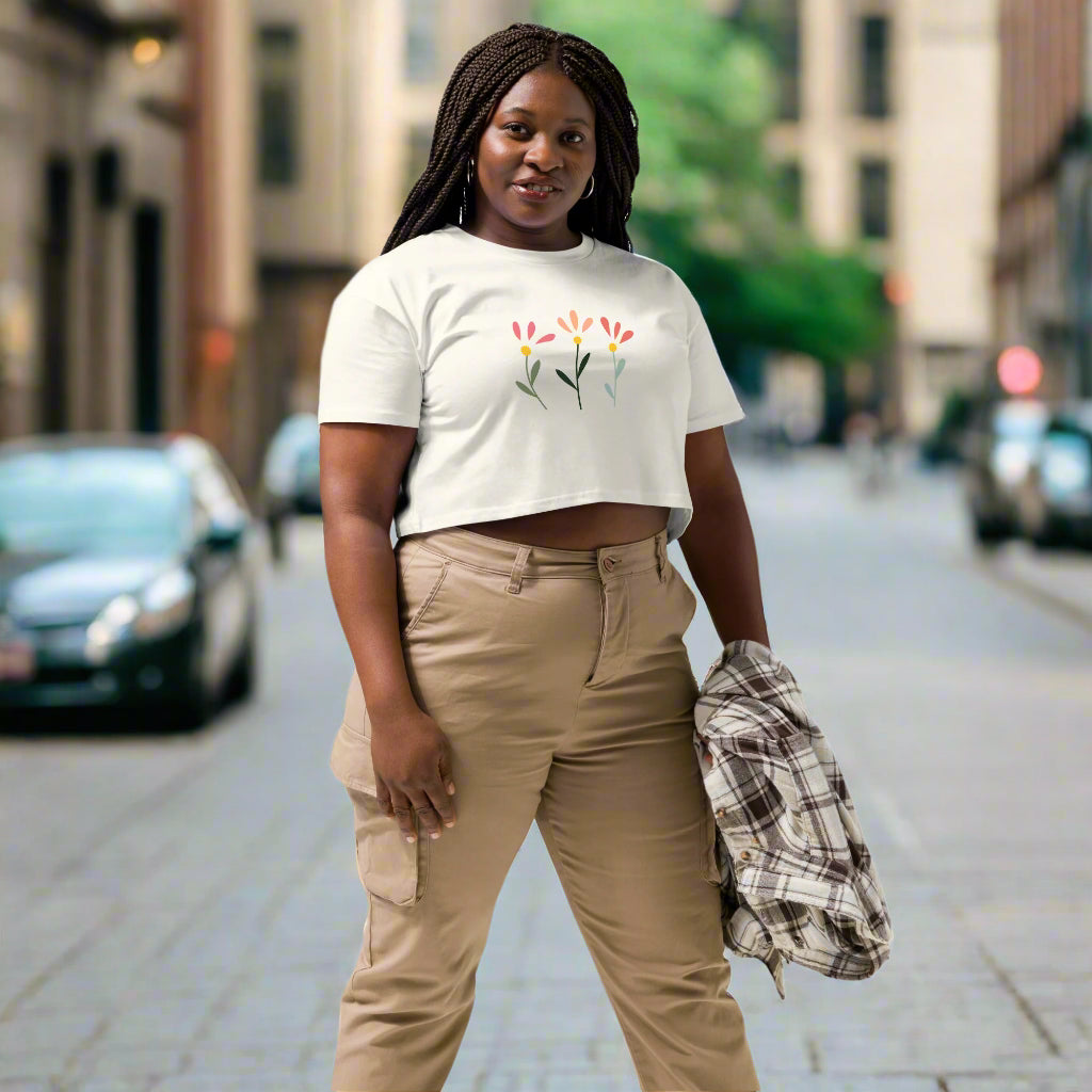 White crop top featuring a delicate printed floral design of three flowers in pink, orange, and yellow with green stems, worn with beige cargo pants and holding a plaid shirt—modern, stylish, and available in multiple colors.