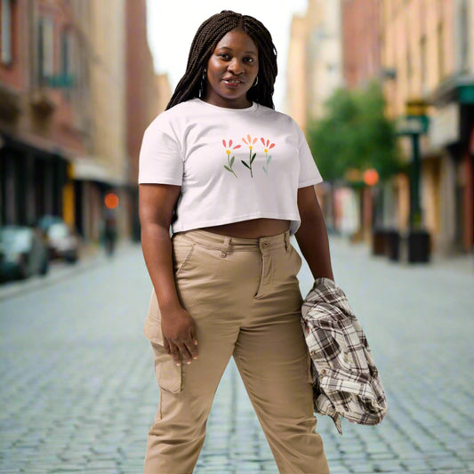 White crop top featuring a delicate printed floral design of three flowers in pink, orange, and yellow with green stems, worn with beige cargo pants and holding a plaid shirt—modern, stylish, and available in multiple colors.